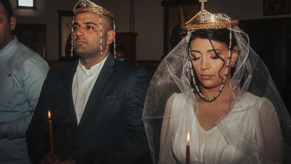 Bride and groom in traditional wedding attire with crowns, holding candles in solemn ceremony.