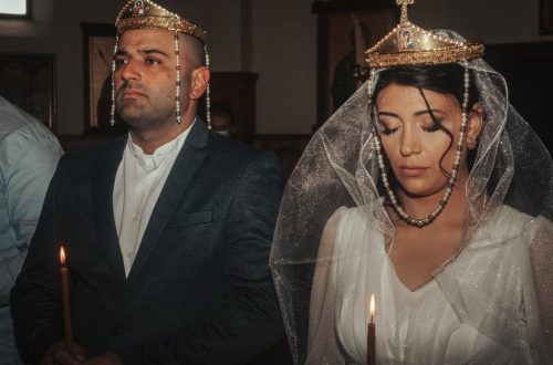 Bride and groom in traditional wedding attire with crowns, holding candles in solemn ceremony.