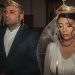 Bride and groom in traditional wedding attire with crowns, holding candles in solemn ceremony.